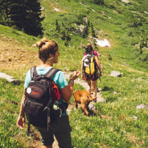 Dos mujeres y un perro caminando por la montaña en un día soleado.